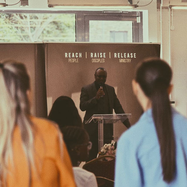 A preacher speaking at the front of a Sunday service beneath the Reach, Raise, Release banner